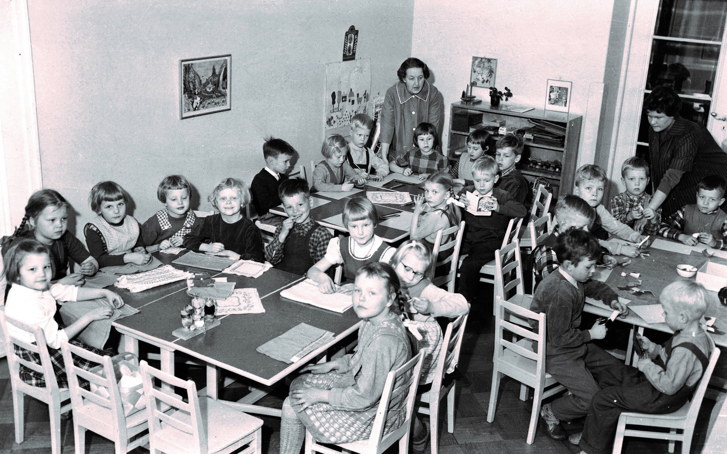 Kuva 2. Ryhmän lapset työskentelevät pöytien ääressä yhdessä huoneessa aikuisten valvonnan alla vuonna 1954. Kuva: Lastentarhamuseon kuvakokoelma.