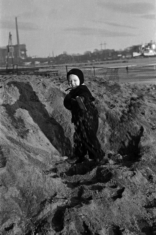 1950 -luku . Lapsi lapioi hiekkaa Sörnäisten rannassa. Taustalla Sörnäisten satamaa.