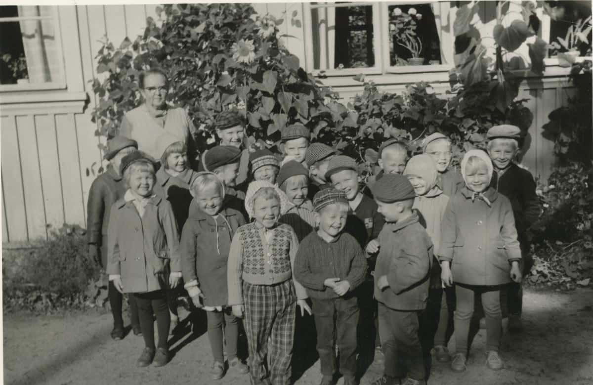 Lastentalon hoitaja 19 lapsen kanssa pihalla kukkaistutusten edessä.; Lastentalon hoitaja lasten kanssa.